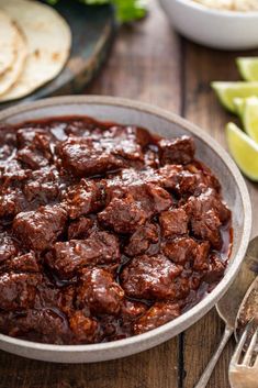 a bowl filled with meat and sauce next to tortilla shells on a wooden table