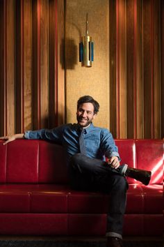 a man sitting on top of a red couch in front of a wooden paneled wall