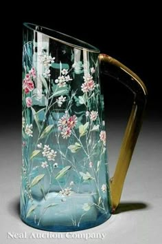 a blue glass pitcher with flowers painted on it's side and gold handle, sitting on a table