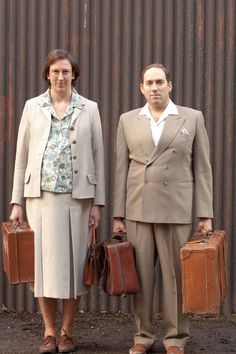 a man and woman standing next to each other with luggage in front of a corrugated wall