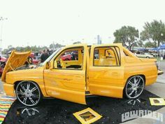 a yellow pick up truck parked in a parking lot with other cars and people around it