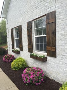 some flowers are in front of a white brick building with shutters on the windows