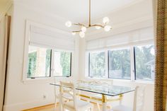 a dining room table with four chairs in front of two windows and a chandelier hanging from the ceiling