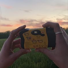 two hands holding up a yellow and black device in front of a field at sunset