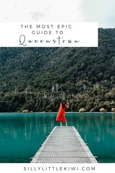 a woman in a red dress standing on the end of a dock with mountains in the background