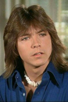 a man with long hair wearing a blue shirt and pearl necklace looking off to the side