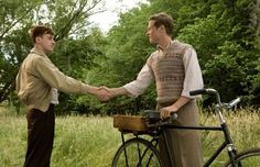 two men shaking hands while standing next to a bike in the grass with trees behind them