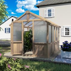 a small wooden greenhouse in front of a white house with blue flowers on the ground