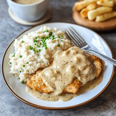 a white plate topped with chicken covered in gravy and mashed potatoes next to a cup of coffee