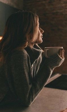 a woman sitting at a table drinking from a coffee cup and looking out the window