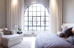 a white bed sitting under a large window next to a chair and ottoman in a bedroom