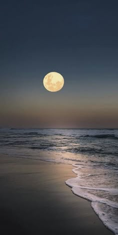 a full moon is seen over the ocean at dusk on an empty beach with waves coming in