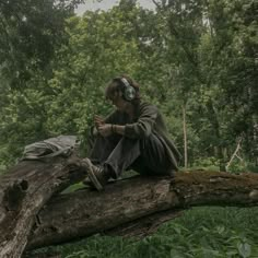 a man sitting on top of a tree branch in the forest using a cell phone