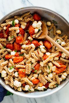 a pan filled with pasta and tomatoes on top of a marble countertop next to a wooden spoon