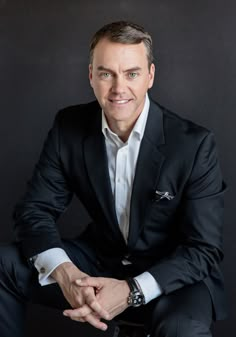 a man in a black suit and white shirt is posing for the camera with his hands on his knees