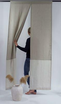 a woman is opening the curtains to look at wheat stalks in a white vase with her feet on the floor