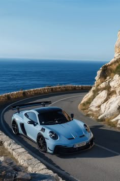 a blue sports car driving down a road next to the ocean on a sunny day