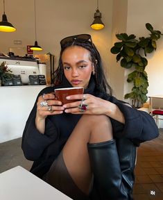 a woman sitting in a chair with her legs crossed and holding a cup while looking at the camera