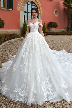 a woman standing in front of a pink building wearing a wedding dress with white flowers on it