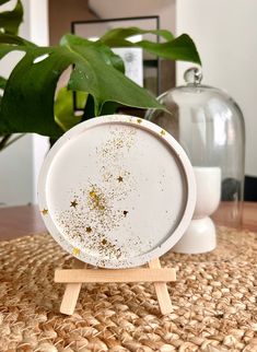 a white plate with gold speckles on it sitting on a wooden stand next to a potted plant