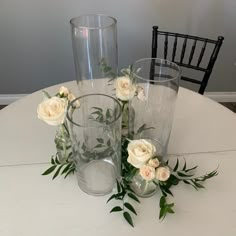 three clear vases with white flowers and greenery sit on a table next to each other