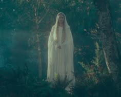 a woman in a white dress and veil standing in the woods with trees behind her