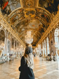 a woman standing in an ornate hall with chandeliers and paintings on the ceiling