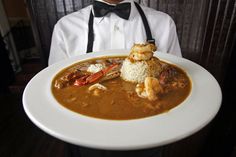 a man in white shirt holding a plate with shrimp and grits on it while wearing an apron