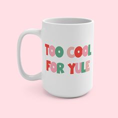 a white coffee mug with the words too cool for yule printed on it, against a pink background