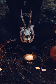 a person sitting on the ground holding a deer skull in front of candles and pumpkins