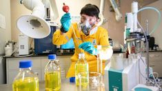 a man in yellow lab coat holding up a beakle filled with liquid and flasks