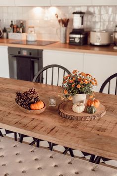 a wooden table topped with two bowls filled with flowers and pumpkins next to a potted plant