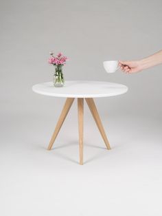 a person holding a coffee cup in front of a white table with wooden legs and a flower vase on top