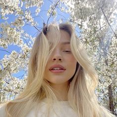 a woman with long blonde hair standing in front of some white flowers and looking up at the sky