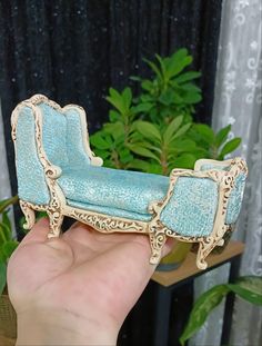 a hand holding a miniature blue couch in front of a potted plant and curtain