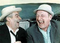 two men laughing while sitting next to each other in front of an old fashioned car