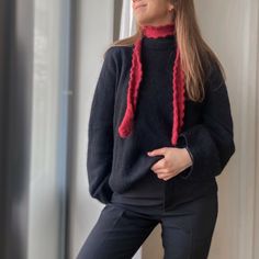 a woman standing in front of a window wearing a black sweater and red knitted scarf