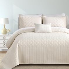 a bed with white comforter and pillows in a room next to a lamp on a dresser