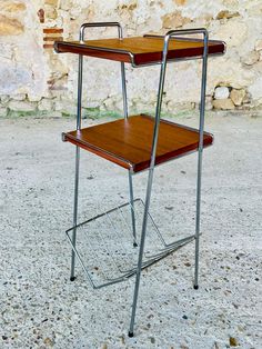 a wooden desk with metal legs and a wood top sitting on the ground in front of a stone wall