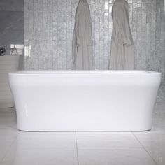 a white bath tub sitting in front of a tiled wall with two robes hanging on it