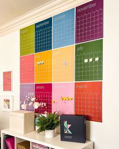 a colorful calendar is mounted on the wall above a white shelf with books and plants