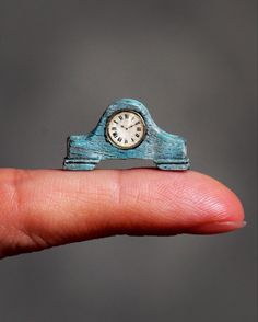 a small blue wooden clock sitting on top of a persons finger in front of a gray background