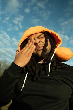 a man with dreadlocks and an orange hoodie covers his face as he talks on the phone