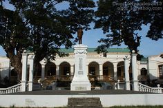 a statue in front of a white building with columns and arches on the sides, surrounded by trees