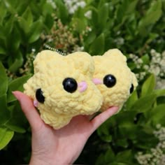 a hand holding two small stuffed animals in front of some bushes and flowers with eyes drawn on them