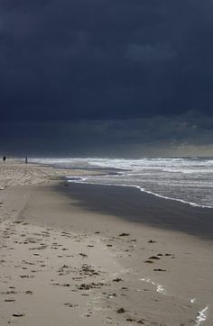 two people are walking along the beach with their surfboards in hand and dark clouds overhead