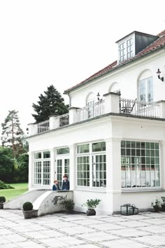 two people sitting on the porch of a large white house with windows and balconies