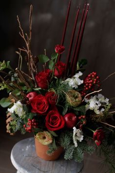 a vase filled with red roses and greenery on top of a wooden table next to candles