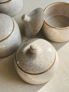 three teapots and two cups sitting on a tableclothed surface, one with speckled glaze