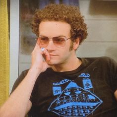 a man with curly hair and glasses talking on a cell phone while sitting in front of a window